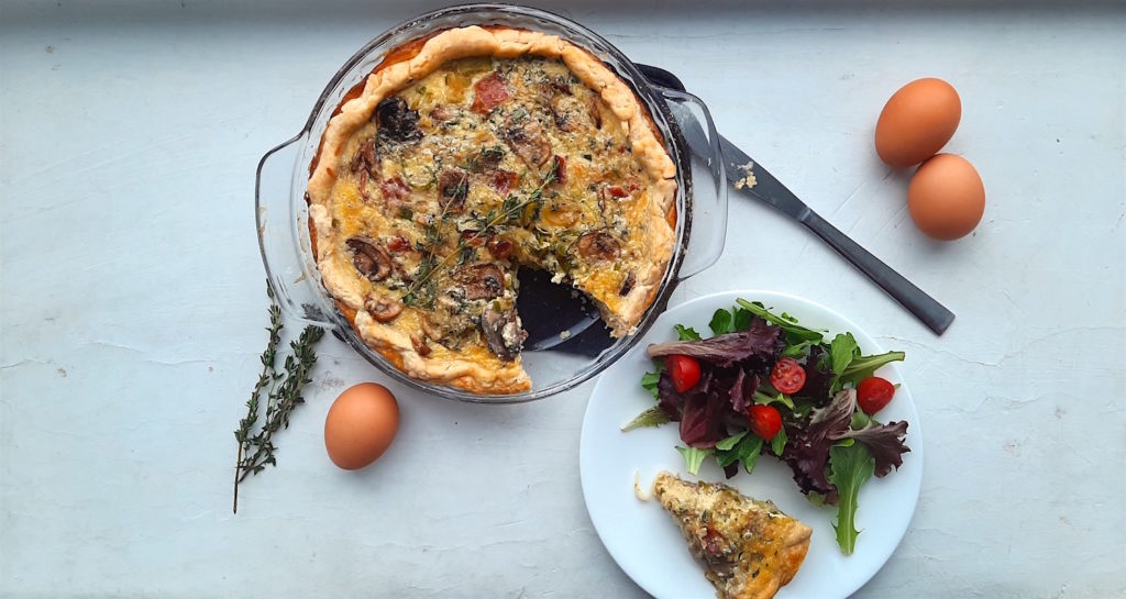 Mushroom, Leek, and Bacon Quiche in a glass pie pan with one slice cut out. Slice is on a white plate with a salad next to the quiche. Surrounded by eggs and thyme stems