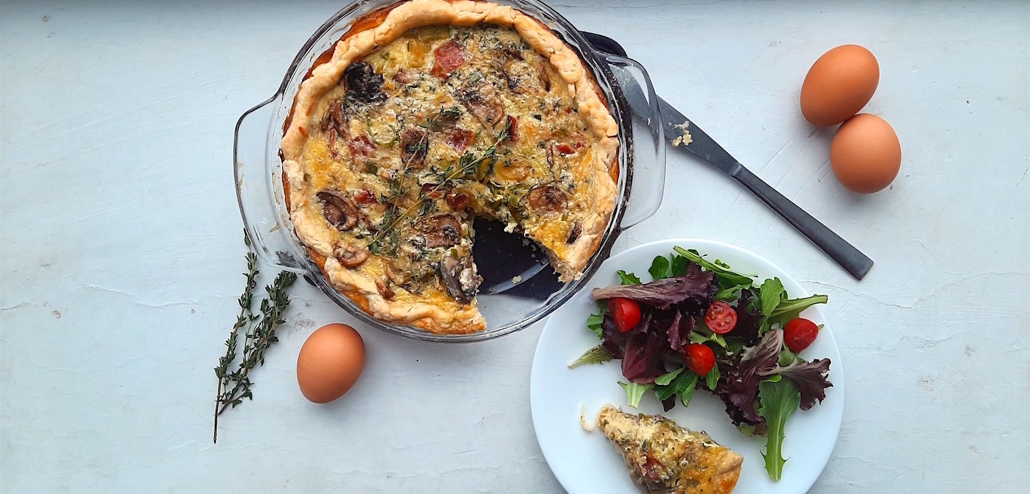 Mushroom, Leek, and Bacon Quiche in a glass pie pan with one slice cut out. Slice is on a white plate with a salad next to the quiche. Surrounded by eggs and thyme stems