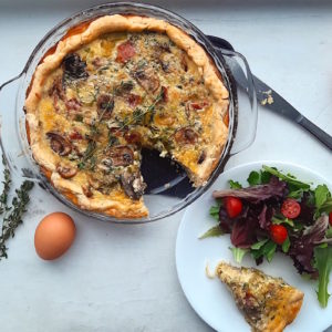 Mushroom, Leek, and Bacon Quiche in a glass pie pan with one slice cut out. Slice is on a white plate with a salad next to the quiche. Surrounded by eggs and thyme stems