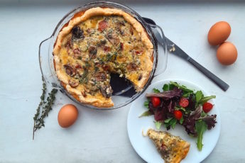 Mushroom, Leek, and Bacon Quiche in a glass pie pan with one slice cut out. Slice is on a white plate with a salad next to the quiche. Surrounded by eggs and thyme stems