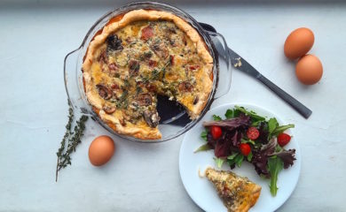 Mushroom, Leek, and Bacon Quiche in a glass pie pan with one slice cut out. Slice is on a white plate with a salad next to the quiche. Surrounded by eggs and thyme stems