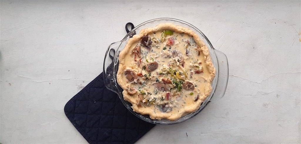 Raw mushroom, leek and bacon quiche in a glass pie dish on a blue oven mit on a white background
