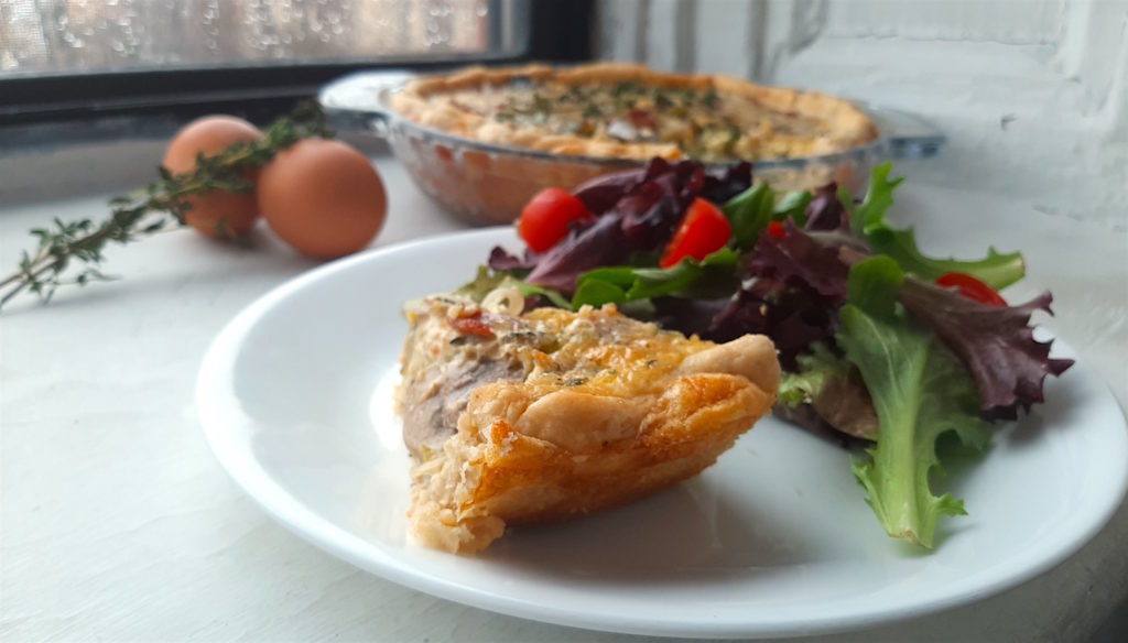 Slice of mushroom, leek and bacon quiche on a white plate with salad in the foreground. Facing away so you can see the crust. Whole mushroom, leek, and bacon quiche in the background in a glass pie dish against a window. Eggs and thyme stems nearby.