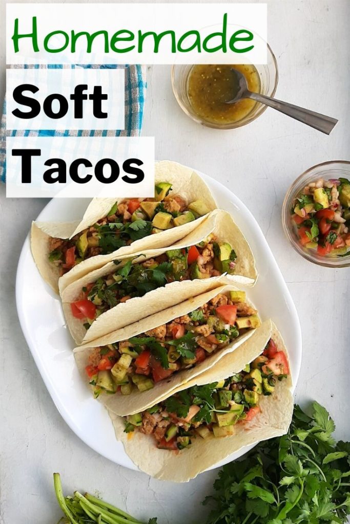 Pinterest image for homemade soft tacos with flour tortillas. Four soft tacos with ground chicken, refried beans, cheddar cheese, shallots, tomatoes, cucumber, avocado, jalapeños, cilantro. Salsa verde and more fresh relish to the side in pyrex bowls.