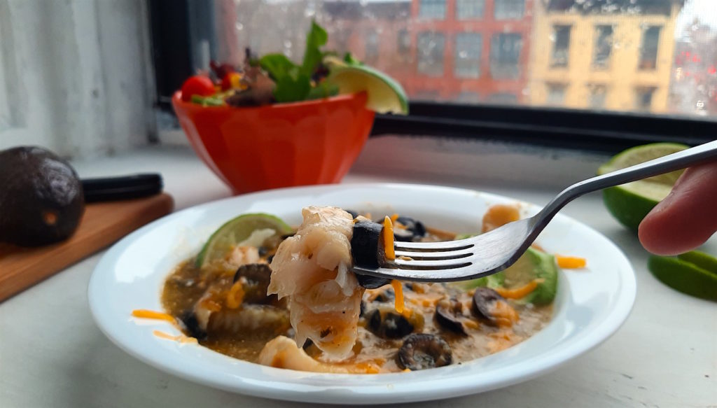 Spicy basa casserole with tomatillo sauce, cheddar cheese, black olives, and lime slices in a white bowl. Woman's hand holding fork above with piece of fish. Surrounded by lime slices, avocado, and taco salad. Sugar with Spice Blog.