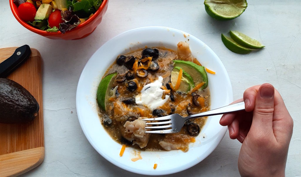 Spicy basa casserole with tomatillo sauce, cheddar cheese, black olives, and lime slices in a white bowl. Woman's hand holding fork above with piece of fish. Surrounded by lime slices, avocado, and taco salad. Sugar with Spice Blog.