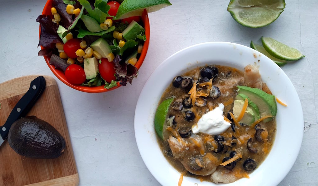 Spicy basa casserole with tomatillo sauce, cheddar cheese, black olives, and lime slices in a white bowl. Woman's hand holding fork above with piece of fish. Surrounded by lime slices, avocado, and taco salad. Sugar with Spice Blog.