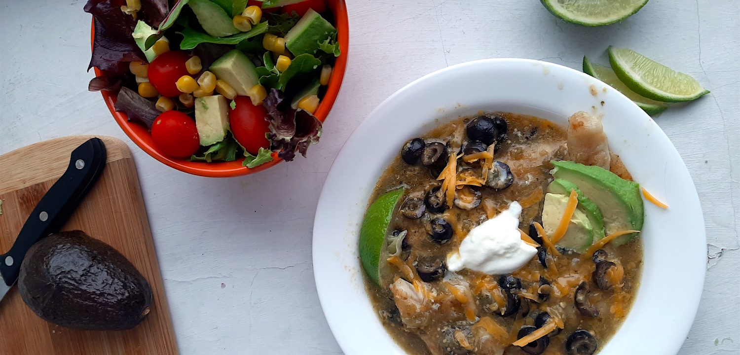 Spicy basa casserole with tomatillo sauce, cheddar cheese, black olives, and lime slices in a white bowl. Woman's hand holding fork above with piece of fish. Surrounded by lime slices, avocado, and taco salad. Sugar with Spice Blog.