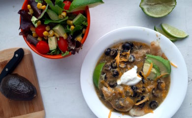 Spicy basa casserole with tomatillo sauce, cheddar cheese, black olives, and lime slices in a white bowl. Woman's hand holding fork above with piece of fish. Surrounded by lime slices, avocado, and taco salad. Sugar with Spice Blog.