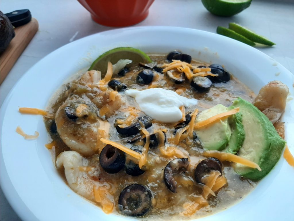 Spicy basa casserole with tomatillo sauce, cheddar cheese, black olives, and lime slices in a white bowl. Surrounded by lime slices, avocado, and taco salad. Sugar with Spice Blog.
