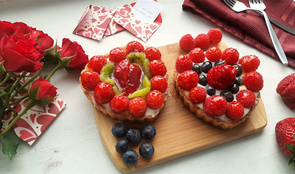 2 Mini heart-shaped fruit tarts with pastry crust, cream cheese filling, and assorted glazed fruit on top. On a wooden cutting board with long stem red roses, raspberries, blue berries, and heart envelopes scattered around