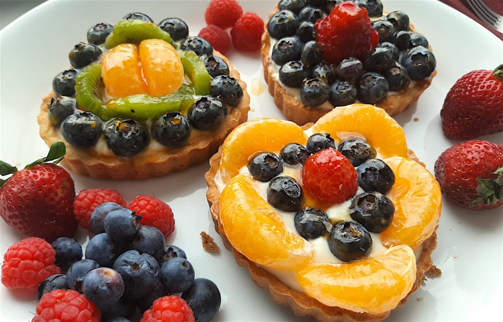 3 Mini heart-shaped fruit tarts with pastry crust, cream cheese filling, and assorted glazed fruit on top. On a white serving tray with strawberries, raspberries, and blueberries scattered around. Sugar with Spice Blog easy mini fruit tarts