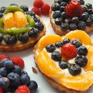 3 Mini heart-shaped fruit tarts with pastry crust, cream cheese filling, and assorted glazed fruit on top. On a white serving tray with strawberries, raspberries, and blueberries scattered around. Sugar with Spice Blog easy mini fruit tarts