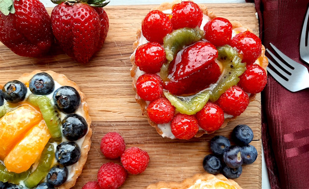 Close up of 2 Mini heart-shaped fruit tarts with pastry crust, cream cheese filling, and assorted glazed fruit on top. On a wooden cutting board with strawberries, raspberries, and blueberries scattered around. Sugar with Spice Blog easy mini fruit tarts