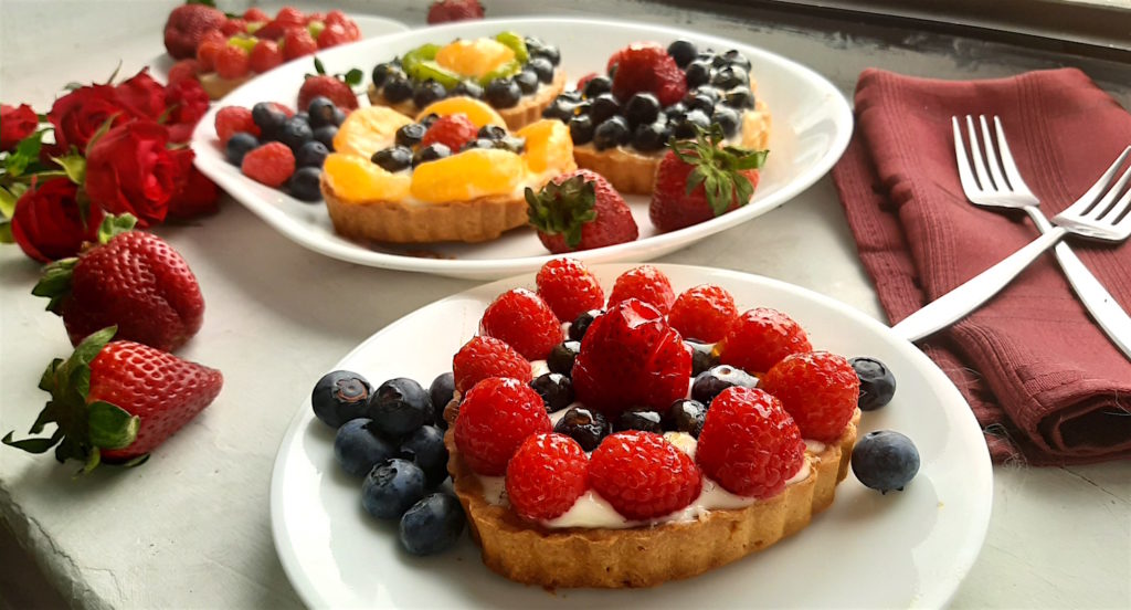 1 Mini heart-shaped fruit tart with pastry crust, cream cheese filling, and assorted glazed fruit on top. On a small white plate, 3 other tarts and assorted fruit in the background. With long stem red roses, blueberries, and red napkins scattered around. Sugar with Spice Blog easy mini fruit tarts