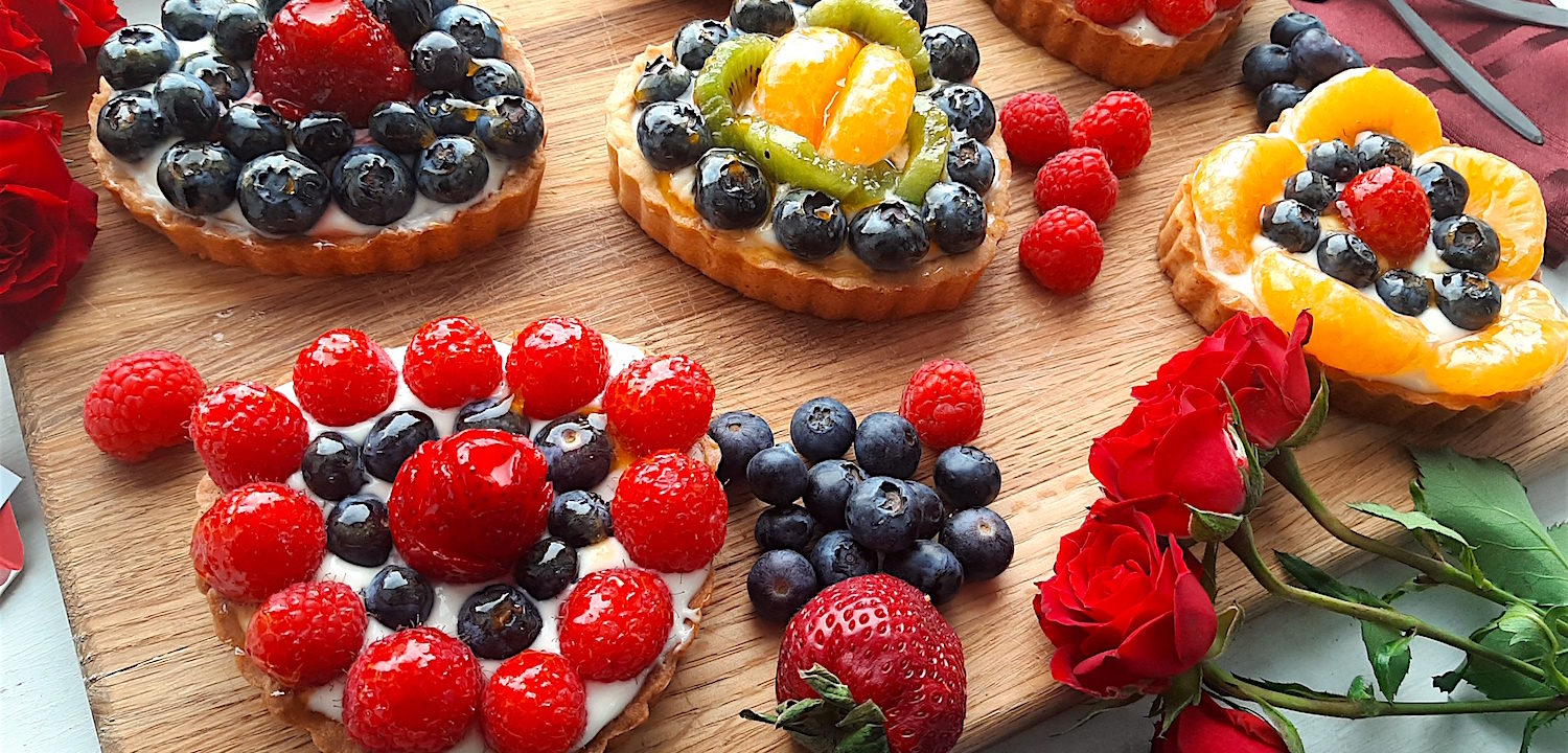 5 Mini heart-shaped fruit tarts with pastry crust, cream cheese filling, and assorted glazed fruit on top. On a wooden cutting board with long stem red roses, raspberries, and blueberries scattered around. Sugar with Spice Blog easy mini fruit tarts
