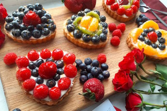 5 Mini heart-shaped fruit tarts with pastry crust, cream cheese filling, and assorted glazed fruit on top. On a wooden cutting board with long stem red roses, raspberries, and blueberries scattered around. Sugar with Spice Blog easy mini fruit tarts