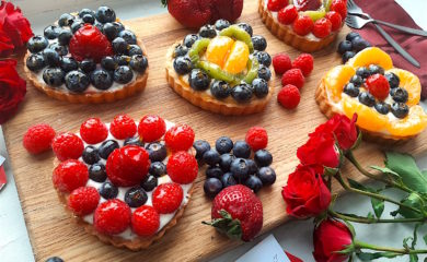 5 Mini heart-shaped fruit tarts with pastry crust, cream cheese filling, and assorted glazed fruit on top. On a wooden cutting board with long stem red roses, raspberries, and blueberries scattered around. Sugar with Spice Blog easy mini fruit tarts