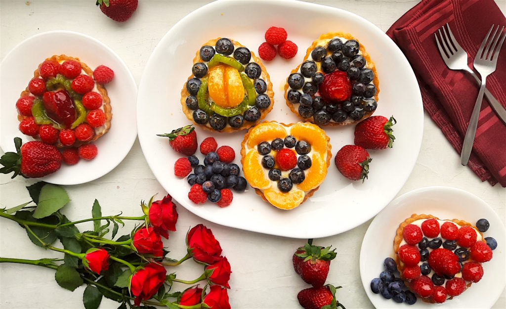 5 Mini heart-shaped fruit tarts with pastry crust, cream cheese filling, and assorted glazed fruit on top. Three tarts on a white serving tray, two tarts on two small white plates with long stem red roses, strawberries, and red napkins scattered around. Sugar with Spice Blog easy mini fruit tarts