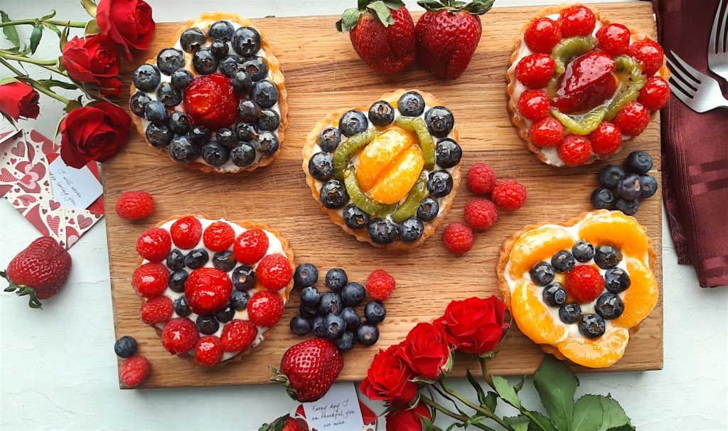 5 Mini heart-shaped fruit tarts with pastry crust, cream cheese filling, and assorted glazed fruit on top. On a wooden cutting board with long stem red roses, raspberries, and blueberries scattered around. Sugar with Spice Blog easy mini fruit tarts