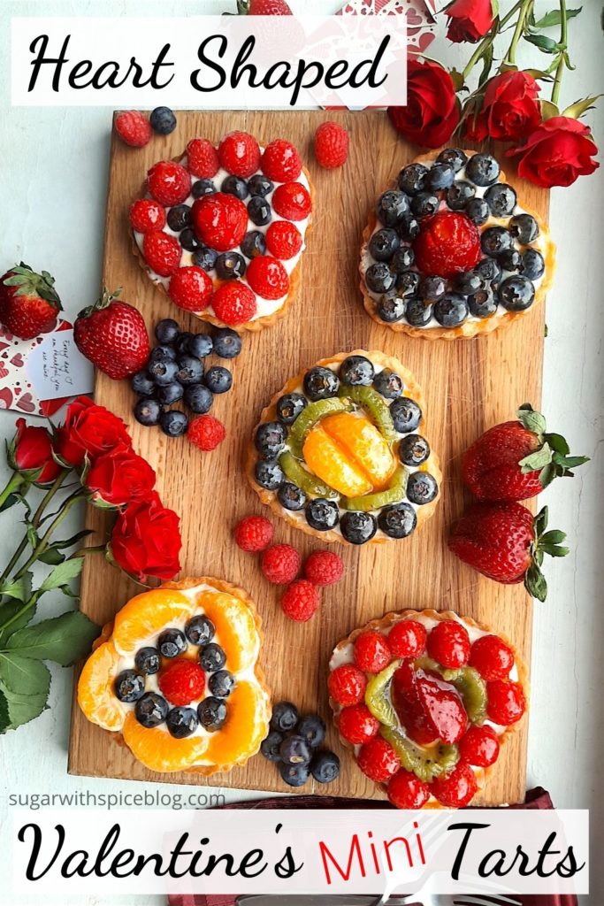 5 Mini heart-shaped fruit tarts with pastry crust, cream cheese filling, and assorted glazed fruit on top. On a wooden cutting board with long stem red roses, raspberries, and blueberries scattered around. Sugar with Spice Blog easy mini fruit tarts. Heart-Shaped Valentine's Mini Fruit Tarts, Pinterest Image