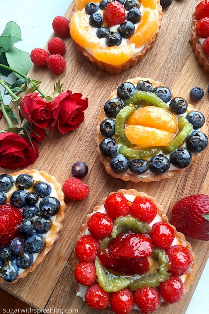 5 Mini heart-shaped fruit tarts with pastry crust, cream cheese filling, and assorted glazed fruit on top. On a wooden cutting board with long stem red roses, raspberries, and blueberries scattered around. Sugar with Spice Blog easy mini fruit tarts