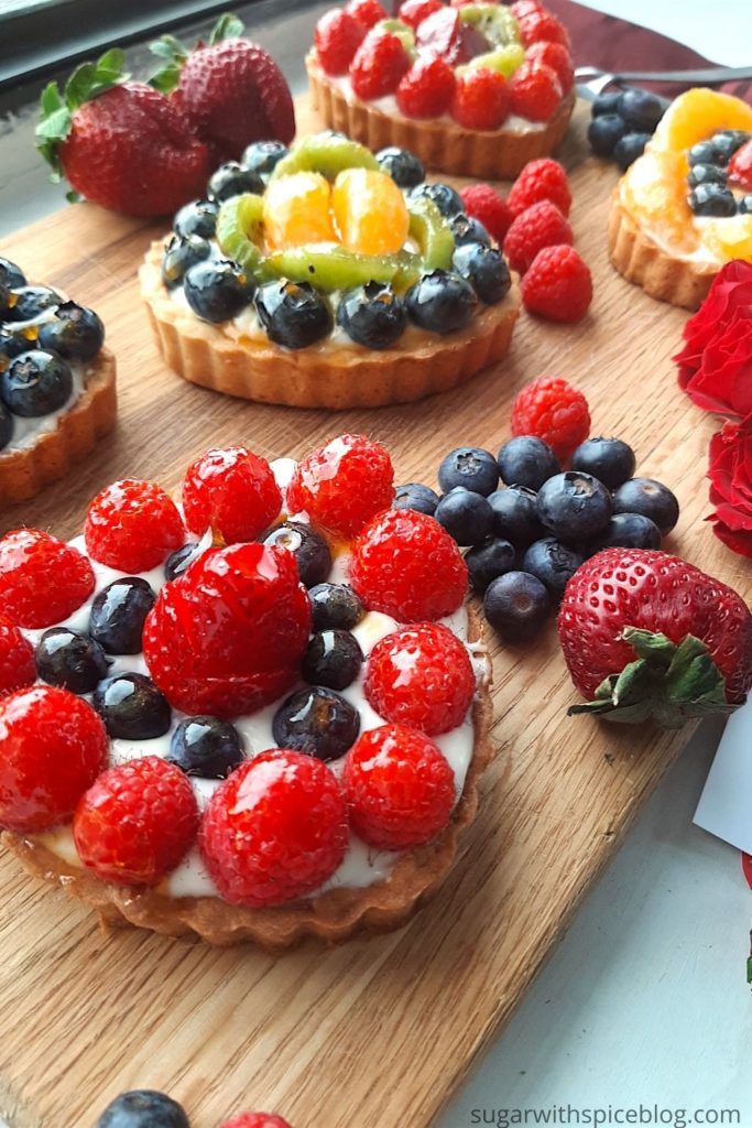 5 Mini heart-shaped fruit tarts with pastry crust, cream cheese filling, and assorted glazed fruit on top. On a wooden cutting board with long stem red roses, raspberries, and blueberries scattered around. Sugar with Spice Blog easy mini fruit tarts