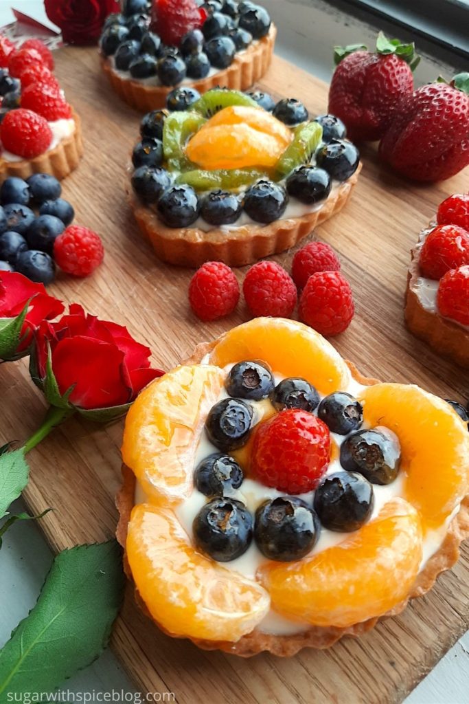 Close up of on mini heart-shaped fruit tart. 5 Mini heart-shaped fruit tarts with pastry crust, cream cheese filling, and assorted glazed fruit on top. On a wooden cutting board with long stem red roses, raspberries, and blueberries scattered around. Sugar with Spice Blog easy mini fruit tarts