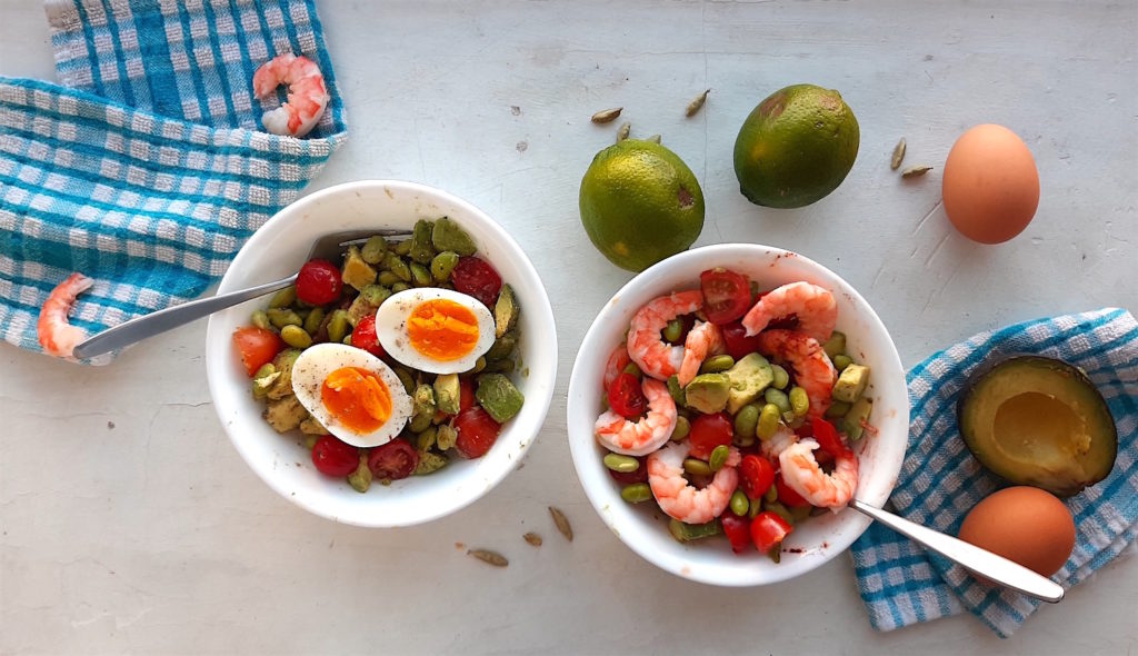 Protein Salad with fresh tomato, avocado, edamame, lime juice, cardamom, shrimp and eggs in white bowls on a white window sill surrounded by blue checked towels and ingredients. Sugar with Spice Blog.