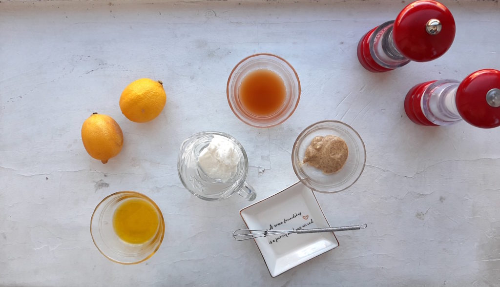 Ingredients for lemony mustard dressing for three bean pasta salad overhead shot on white background. Two lemons, apple cider vinegar in a pyrex ramekin, mustard in a pyrex ramekin, olive oil in a pyrex ramekin, yogurt in a glass dressing holder, red salt and pepper shakers