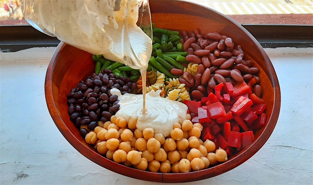 Lemony Mustard Dressing being poured on Three Bean Pasta Salad ingredients. Tri-color rotini, chickpeas, chopped red bell pepper, kidney beans, cooked green beans, black beans in a wooden salad bowl on a window sill.