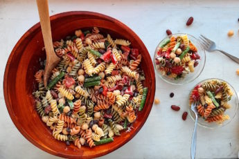 Three Bean Pasta Salad with lemony mustard dressing Sugar with Spice Blog. Overhead shot of Three Bean Pasta Salad in a big wooden salad bowl and wooden spoon with two small pyrex ramekins also holding salad on the right with two silver forks and some dish towels on a white surface.