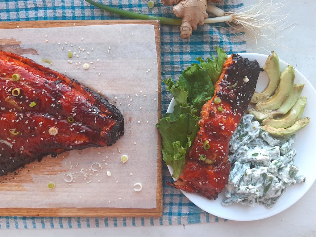 Slice of honey wasabi salmon on a white plate with lettuce garnish, yogurt dill green beans, avocado slices on a white plate. Next to the tail end of wasabi honey glazed salmon on a wooden cutting board surrounded by fresh ginger, scallions, and blue checked dish towel. Sugar with Spice Blog.