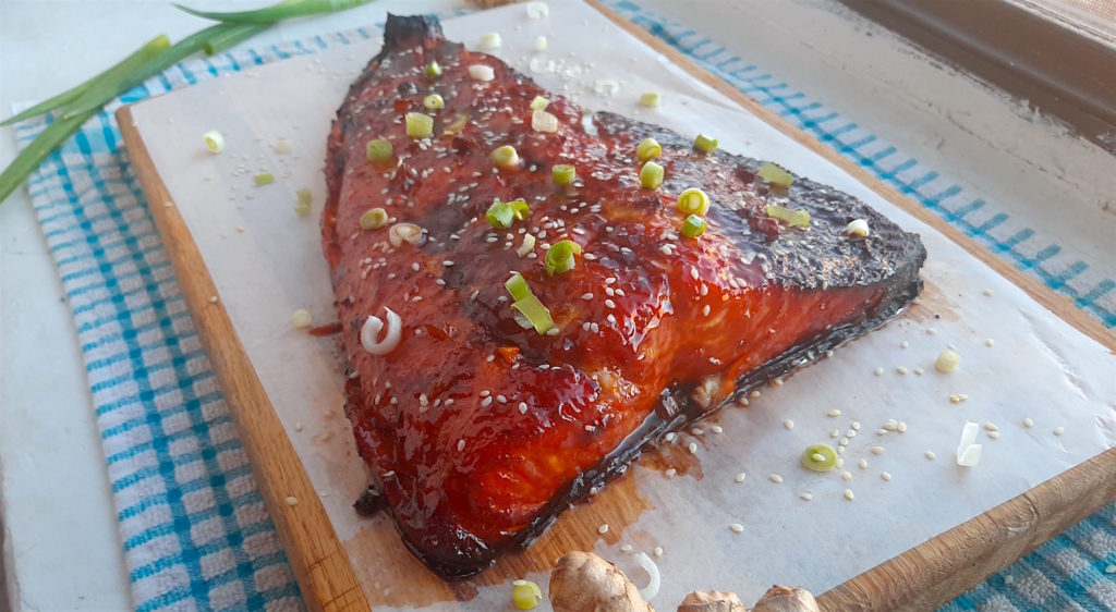Honey Wasabi Glazed Salmon face on, resting on a wooden cutting board and blue checked dish towel with scallions, fresh ginger, and sesame seeds. Sugar with Spice Blog.