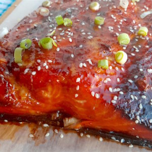 Honey and Wasabi Glazed Salmon close up topped with sesame seeds and sliced green scallions on a wooden cutting board with blue checked dish towel in the background.