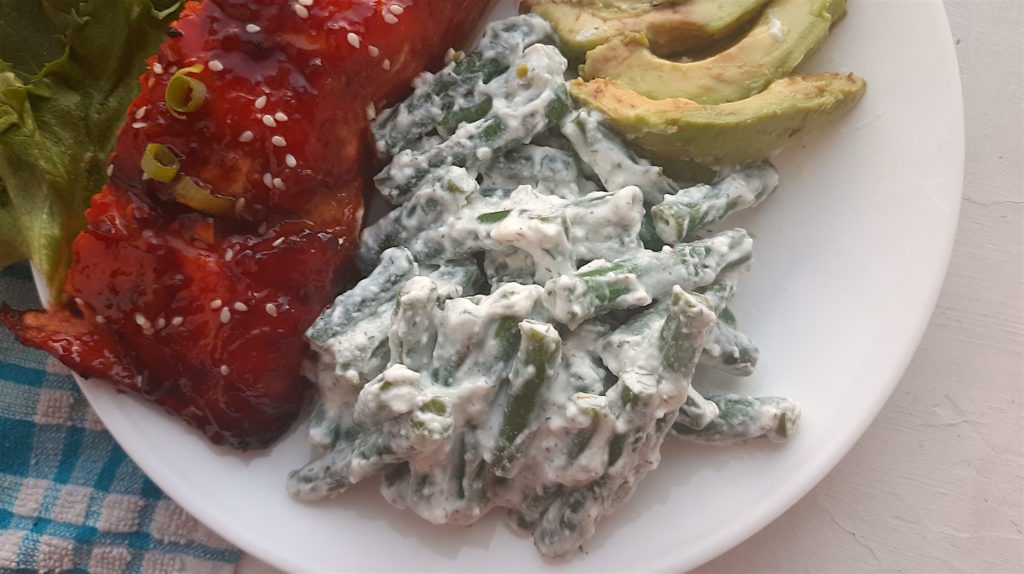 Close up of green beans in yogurt and dill sauce on a white plate with avocado slices and baked salmon in the background. Sugar with Spice Blog.