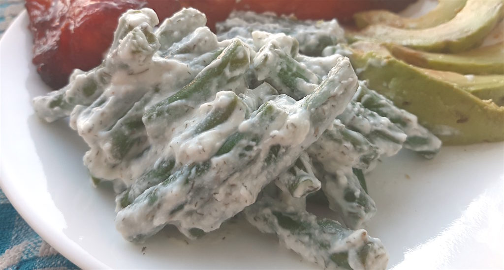 Close up of green beans in yogurt and dill sauce on a white plate with avocado slices and baked salmon in the background. Sugar with Spice Blog.