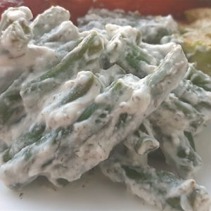 Close up of green beans in yogurt and dill sauce on a white plate with avocado slices and baked salmon in the background.