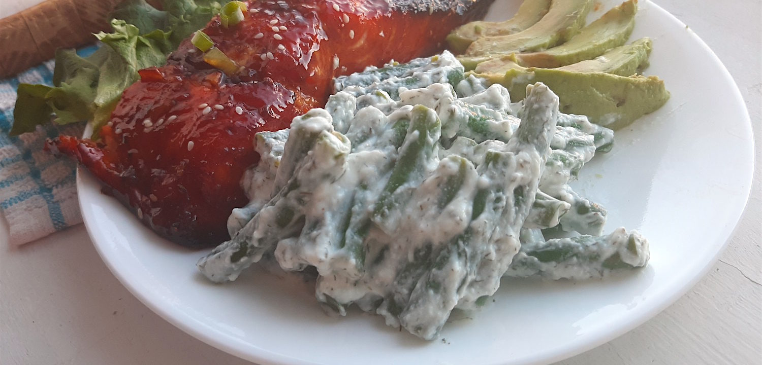 Close up of green beans in yogurt and dill sauce on a white plate with avocado slices and baked salmon in the background.