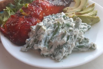 Close up of green beans in yogurt and dill sauce on a white plate with avocado slices and baked salmon in the background.