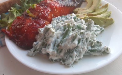 Close up of green beans in yogurt and dill sauce on a white plate with avocado slices and baked salmon in the background.