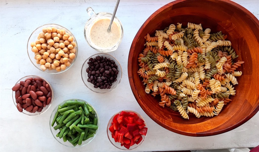 Overhead shot of three bean pasta salad ingredients. Cooked tri-color rotini pasta in wooden salad bowl with wooden spoon, lemony mustard dressing in glass serving dish with silver whisk, chickpeas in glass ramekin, kidney beans in glass ramekin, green beans in glass ramekin, chopped red bell pepper in glass ramekin, black beans in glass ramekin on white background. Sugar with Spice Blog.