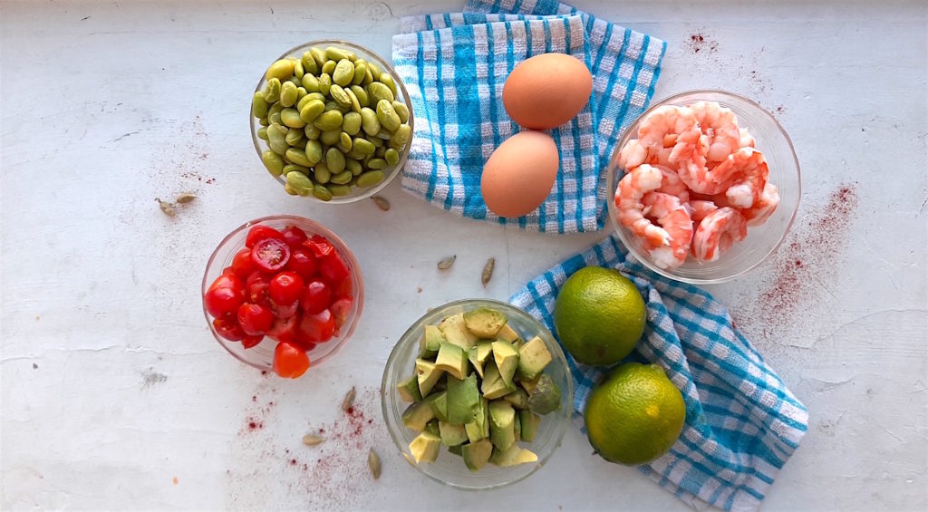 Ingredients for protein salad. Edamame, sliced cherry tomatoes, sliced avocado, limes, cardamom seeds, steamed shrimp, and eggs on a white background with blue checked towels. Sugar with Spice Blog.