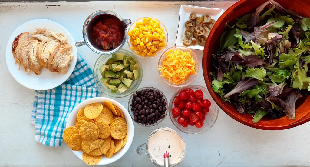 Spicy Santa Fe Taco Salad ingredients. Salad greens in a wooden bowl with sliced chicken, tortilla chips, black beans, tomatoes, corn, avocado, cheddar cheese, salsa, and creamy cilantro ranch and a blue dishtowel on a white window sill