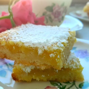 Close up of 2 Lemon Love Note bars on a tea saucer with tea cup, second plate of lemon love notes and pink spray roses in the background.