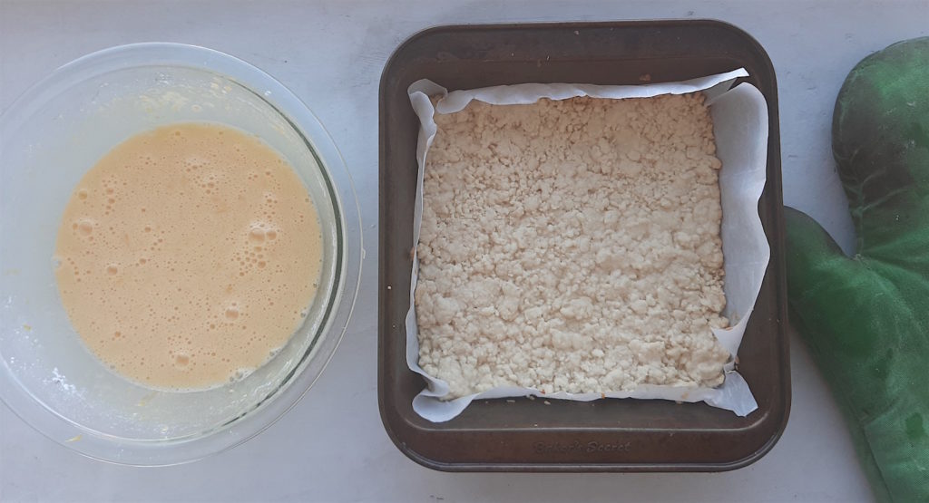Freshly baked shortbread crust for lemon love notes with pyrex bowl of lemon filling, green oven mitt on a white background.