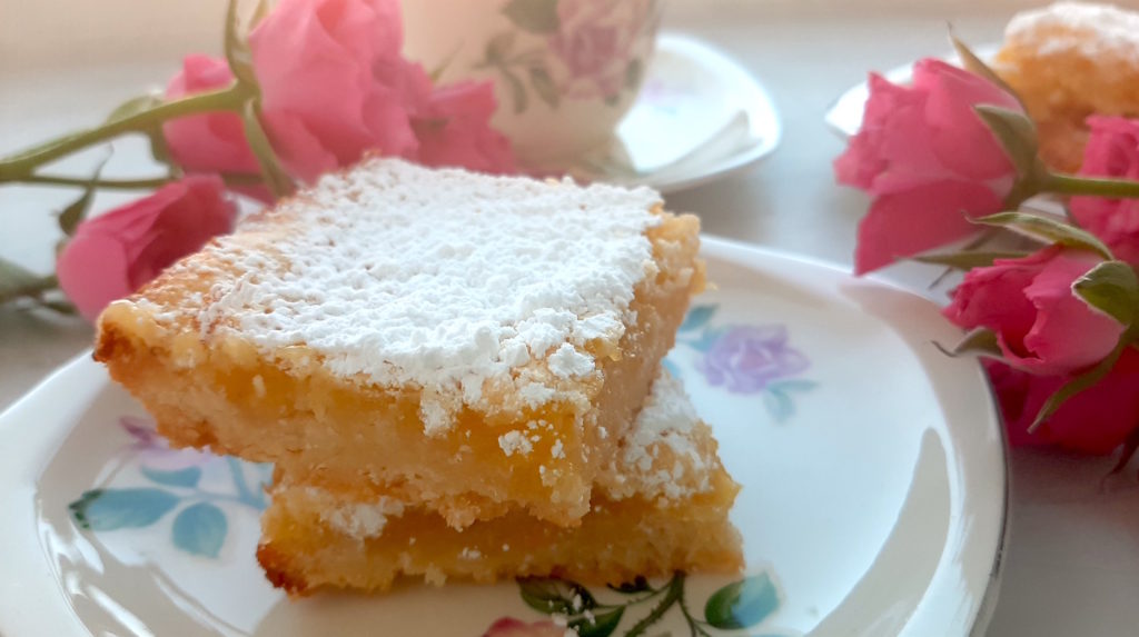 Close up of 2 Lemon Love Note bars on a tea saucer with tea cup, second plate of lemon love notes and pink spray roses in the background.