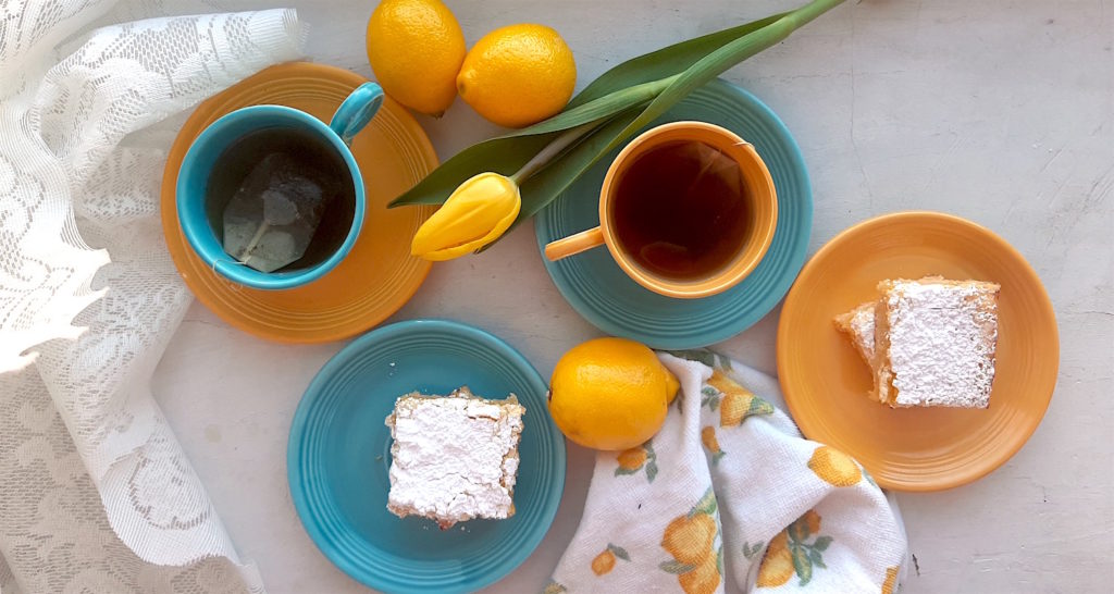 Lemon Love Note lemon cookie bars on blue and yellow tea saucers with two cups of tea in mismatched blue and yellow cups and saucers surrounded by lemons, yellow tulips, lace, and lemon dishtowels. Sugar with Spice Blog.