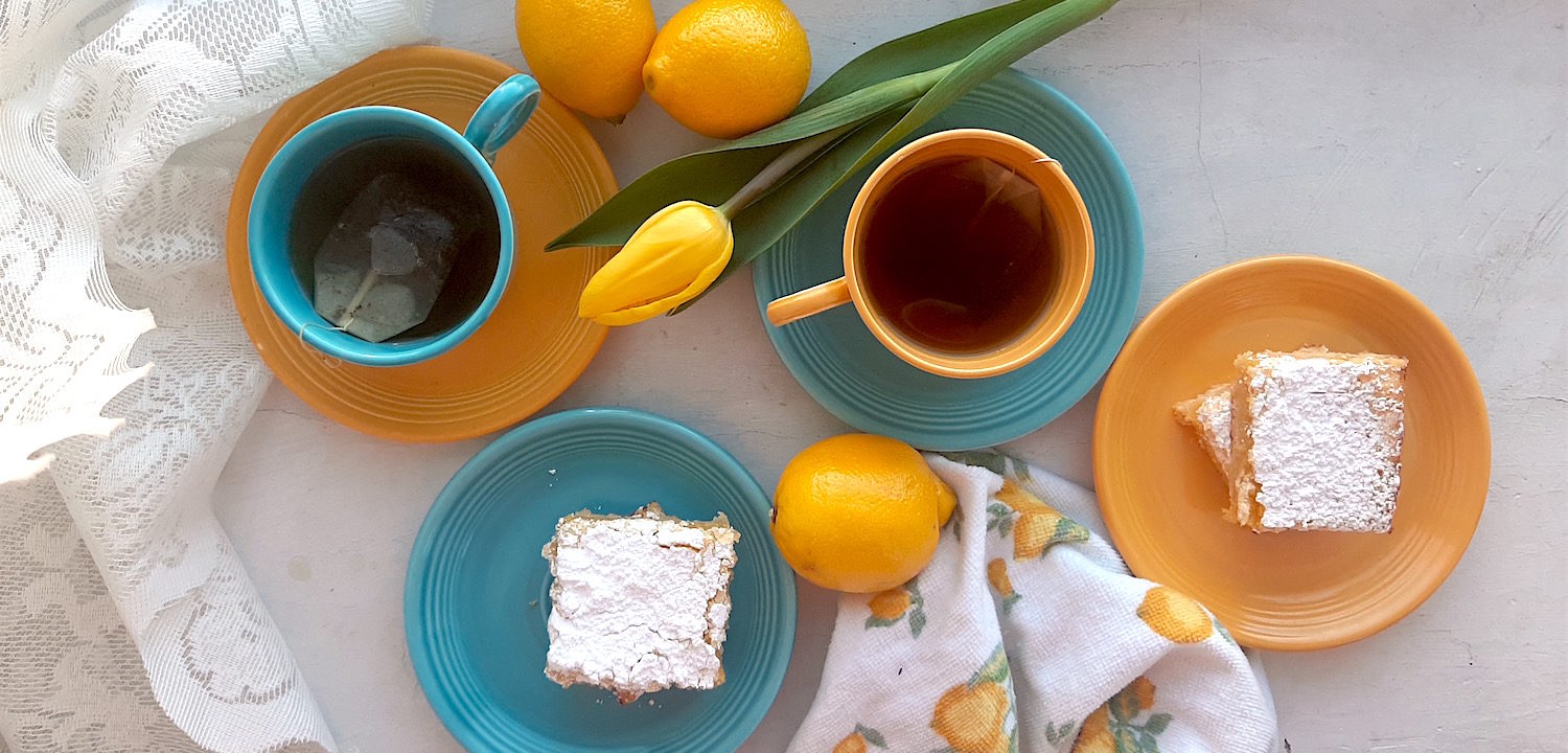 Lemon Love Note lemon cookie bars on blue and yellow tea saucers with two cups of tea in mismatched blue and yellow cups and saucers surrounded by lemons, yellow tulips, lace, and lemon dishtowels. Sugar with Spice Blog.