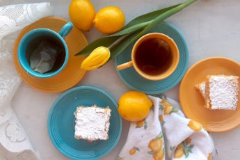 Lemon Love Note lemon cookie bars on blue and yellow tea saucers with two cups of tea in mismatched blue and yellow cups and saucers surrounded by lemons, yellow tulips, lace, and lemon dishtowels. Sugar with Spice Blog.